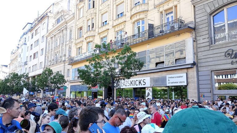 Pride marchers gathered along a Budapest street lined with ornate building facades and shopfronts