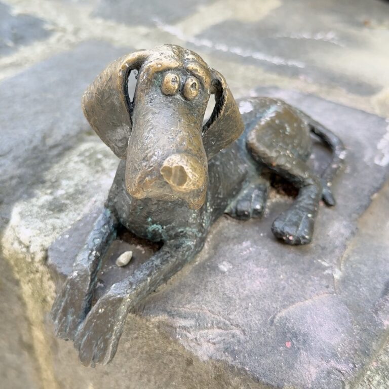 A small bronze dog lying flat on a stone block.