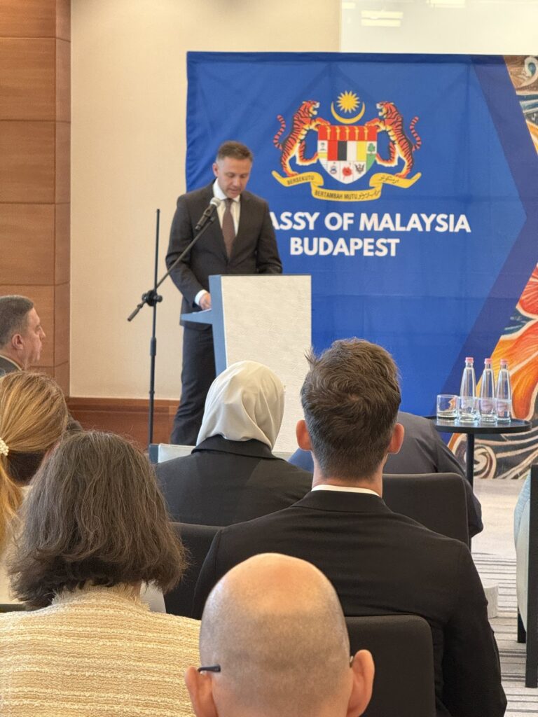 Áron Littvay-Kovács speaking at a lectern in front of a blue banner reading Embassy of Malaysia Budapest.