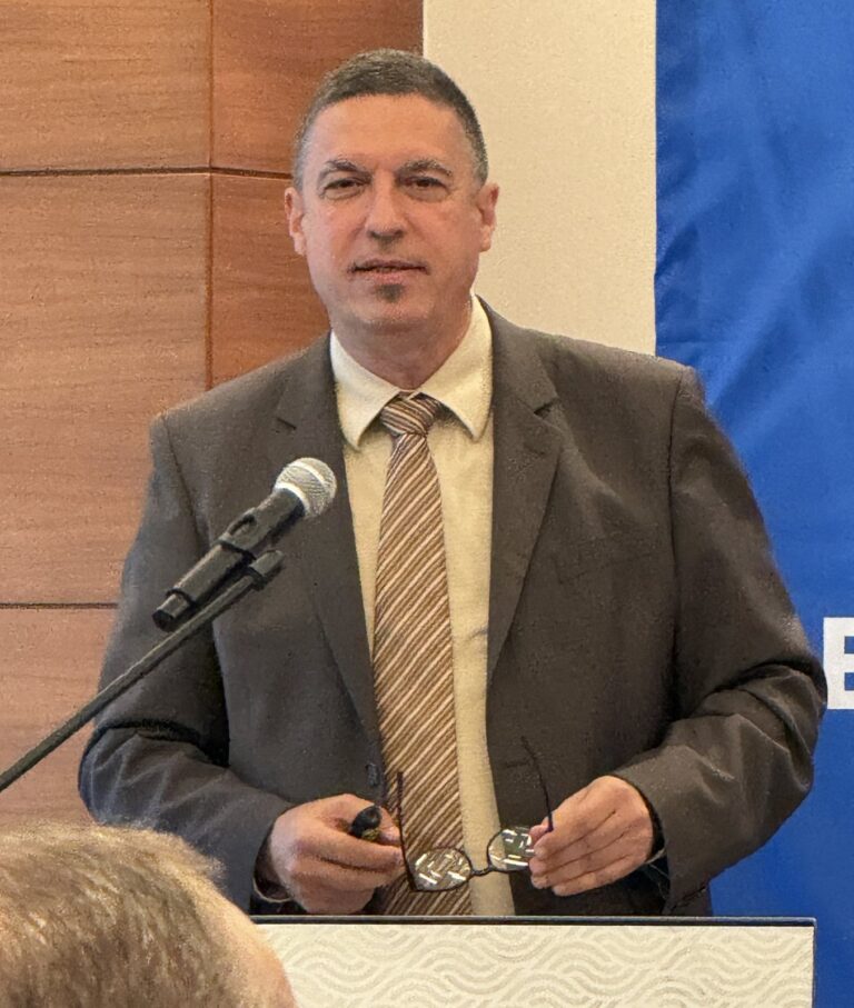 Zoltán Kiss speaking into a microphone at a lectern in front of a blue banner.