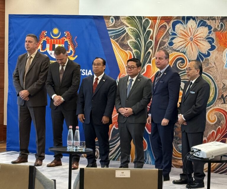 Six men standing on a stage in front of a blue Malaysian coat of arms backdrop at a business forum in Budapest.